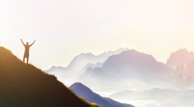 Man on peak of mountain. Emotional scene. Young man with backpack standing with raised hands on top of a mountain and enjoying mountain view. Hiker on the mountain top. Sport and active life concept.